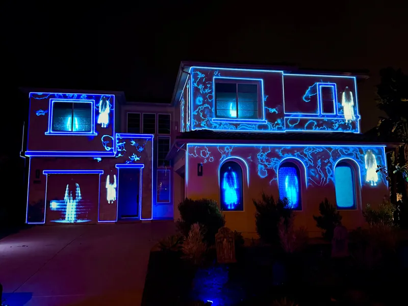 Spectral figures projected in the windows and on the garage of a blue-outlined house facade, Halloween 2025