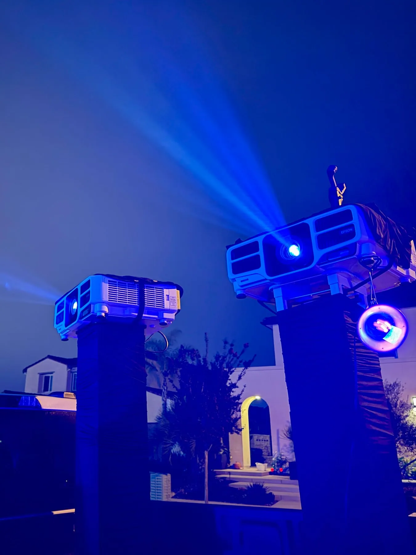 Projectors on pillars beaming blue light toward a house at night, Halloween 2025