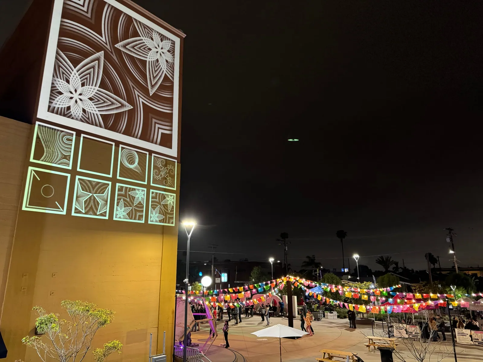 Projected floral mural panels on a building facade beside a festival plaza at night